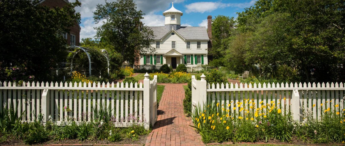 Straight-on shot of exterior of Cupola House, with white picket fence and gardens in front of house during cloudy daytime