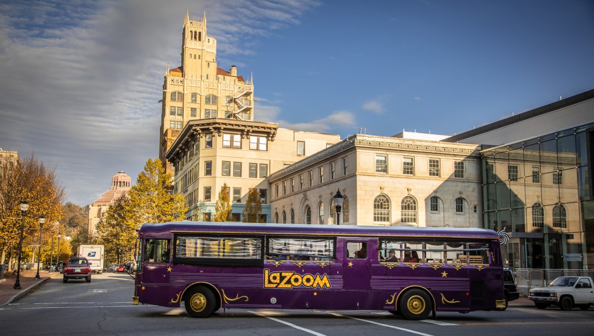 Purple LaZoom comedy bus driving through downtown Asheville during daytime.