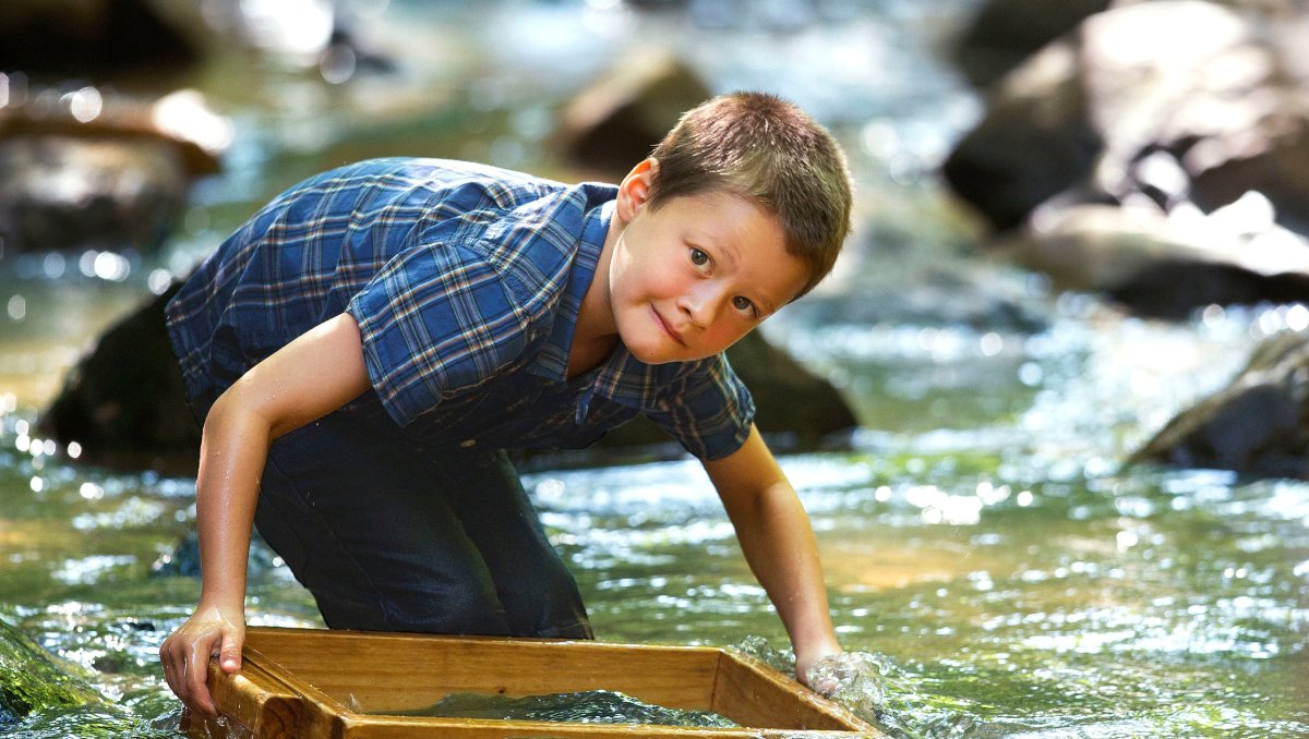 A young boy kneels in a shallow, rocky stream, using a wooden sifting box to pan for treasures in the clear water. Dappled sunlight filters through the trees, illuminating the serene, nature-filled scene.