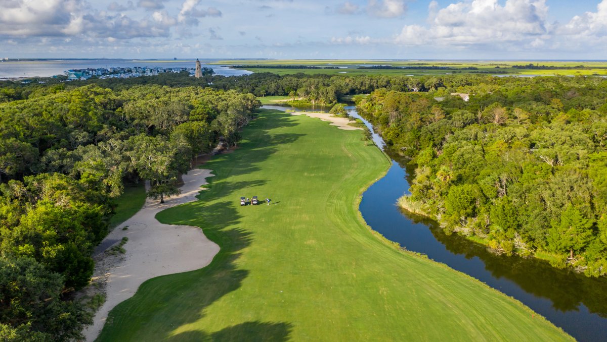 Aerial of golf course surrounded by stream, trees and marshes in background