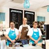 Three coffee shop workers posing behind register with coffee menu on wall behind them.