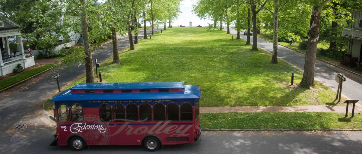 Horizontal shot of tree-lined green lawn in front of historic courthouse with Edenton Trolley parked in front during daytime