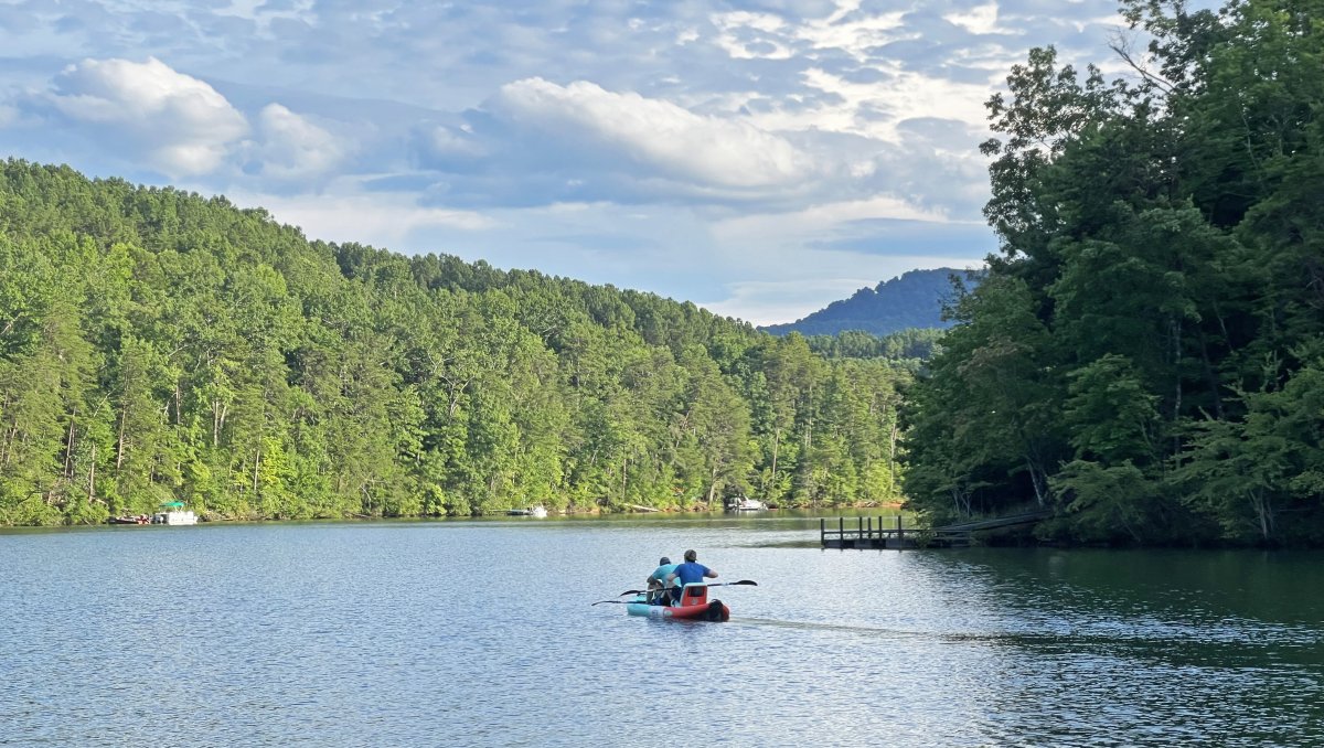 Two people in kayak in middle of lake surrounded by trees and small mountains.