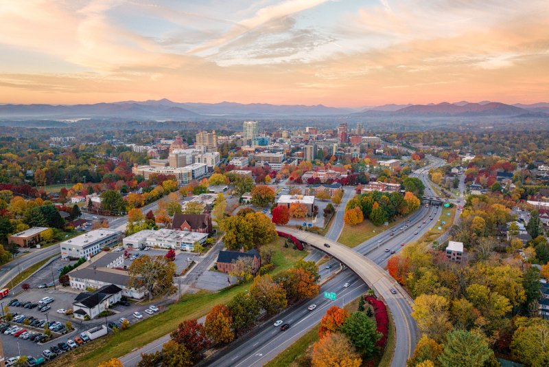 Aerial view of Asheville’s city skyline in fall, surrounded by colorful autumn foliage. Roads curve through the cityscape, with mountains in the distance and a warm sunset sky overhead.