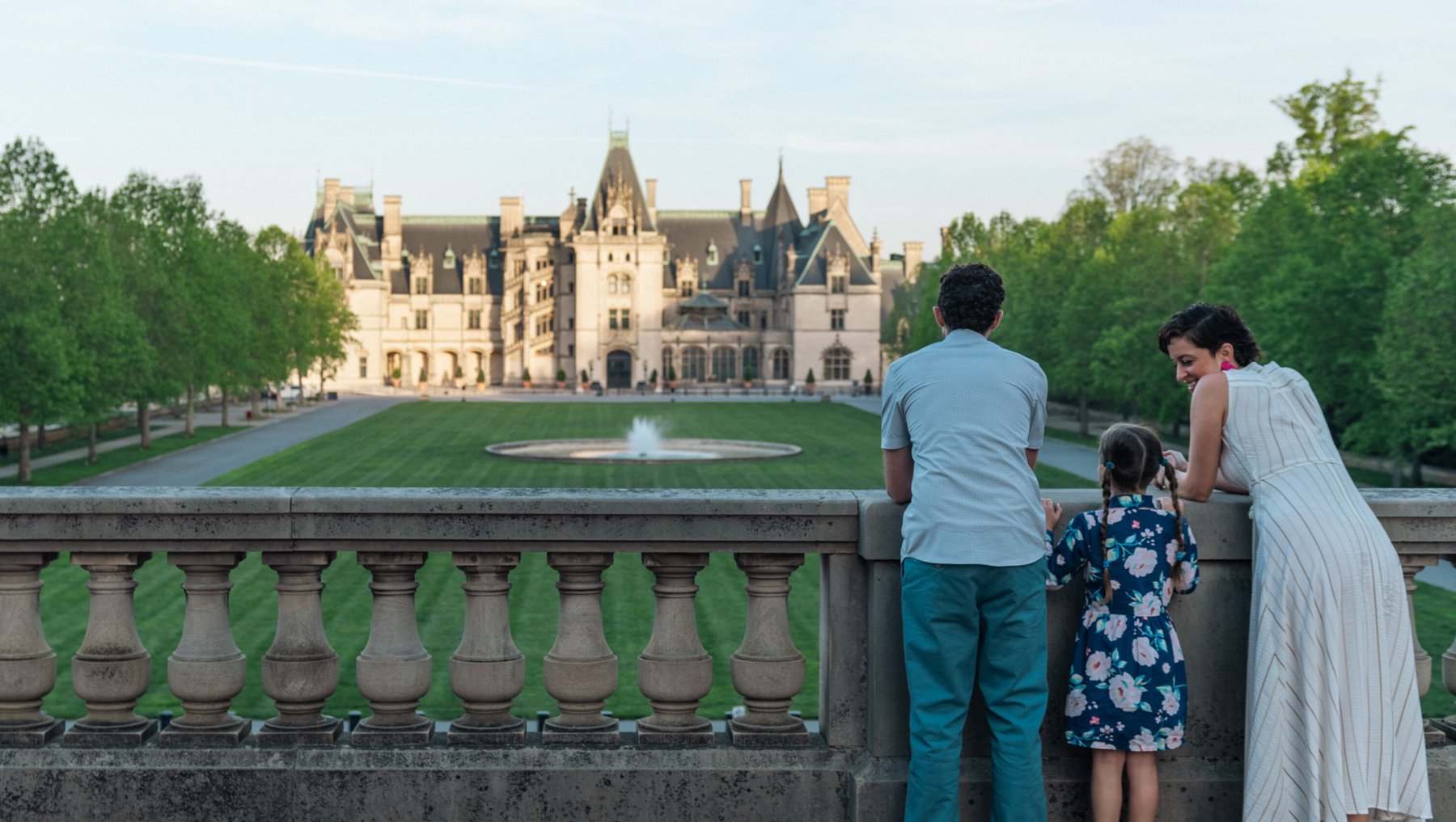 Parents and child with backs to us standing on ornate walkway overlooking Biltmore house and grounds