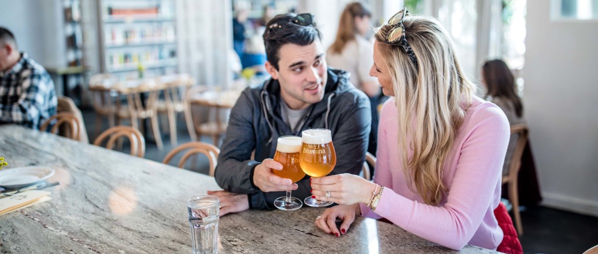 Couple cheersing beer at bar with whimsical lights and bookshelves behind them in restaurant