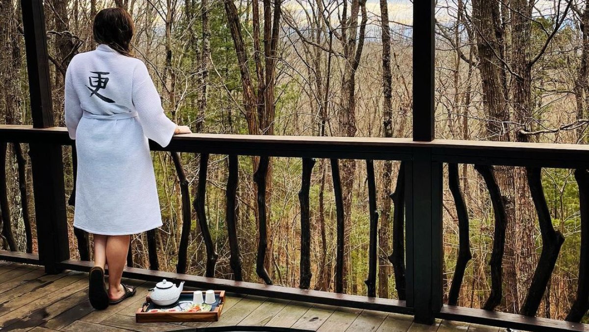 Person in robe standing outside soaking tub looking into forest at spa.