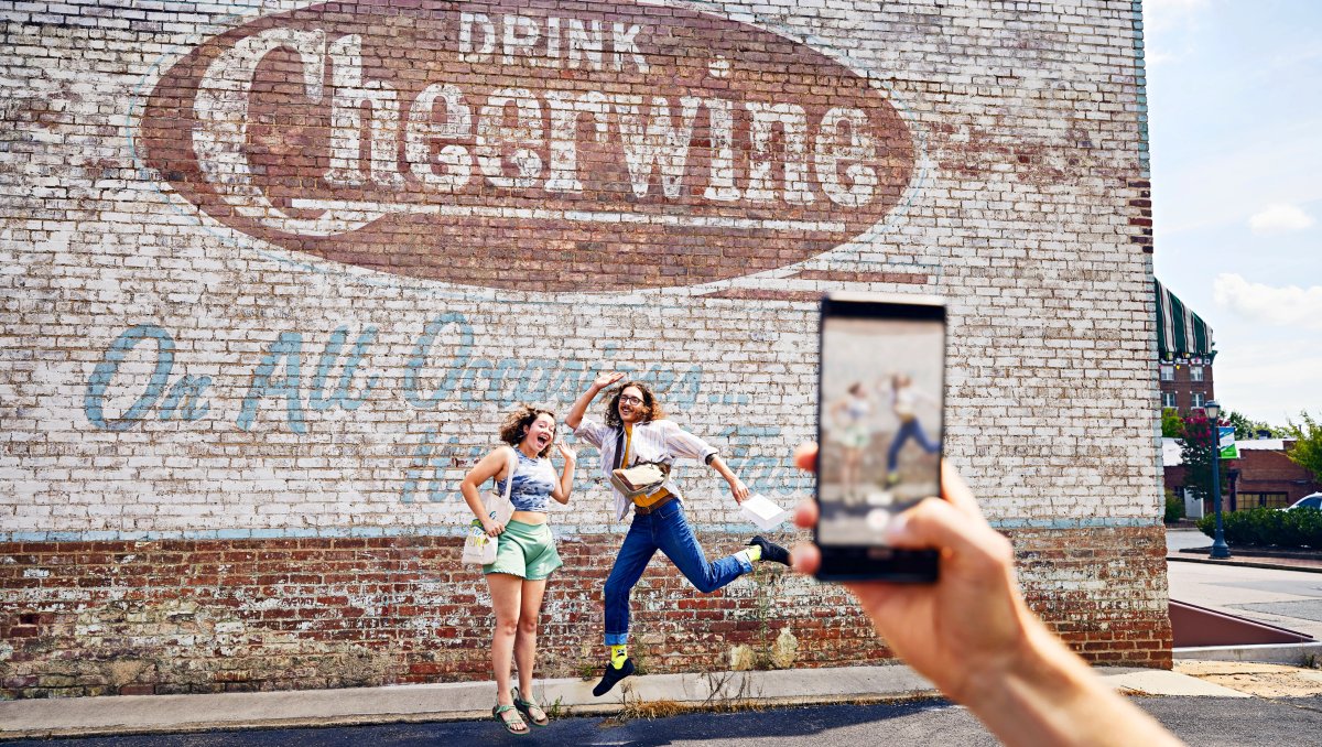 Phone in foreground taking photo of friends jumping in front of Cheerwine mural on side of building