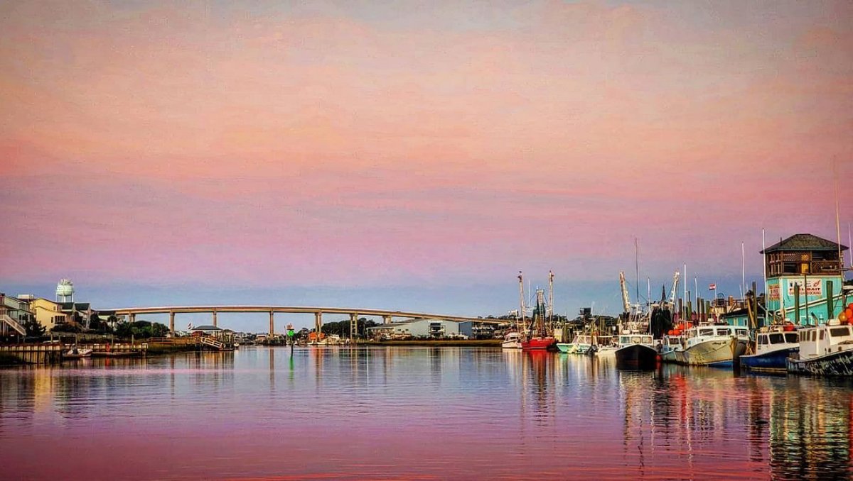 Waterway with boats docked on side and bridge in distance under a pink sky