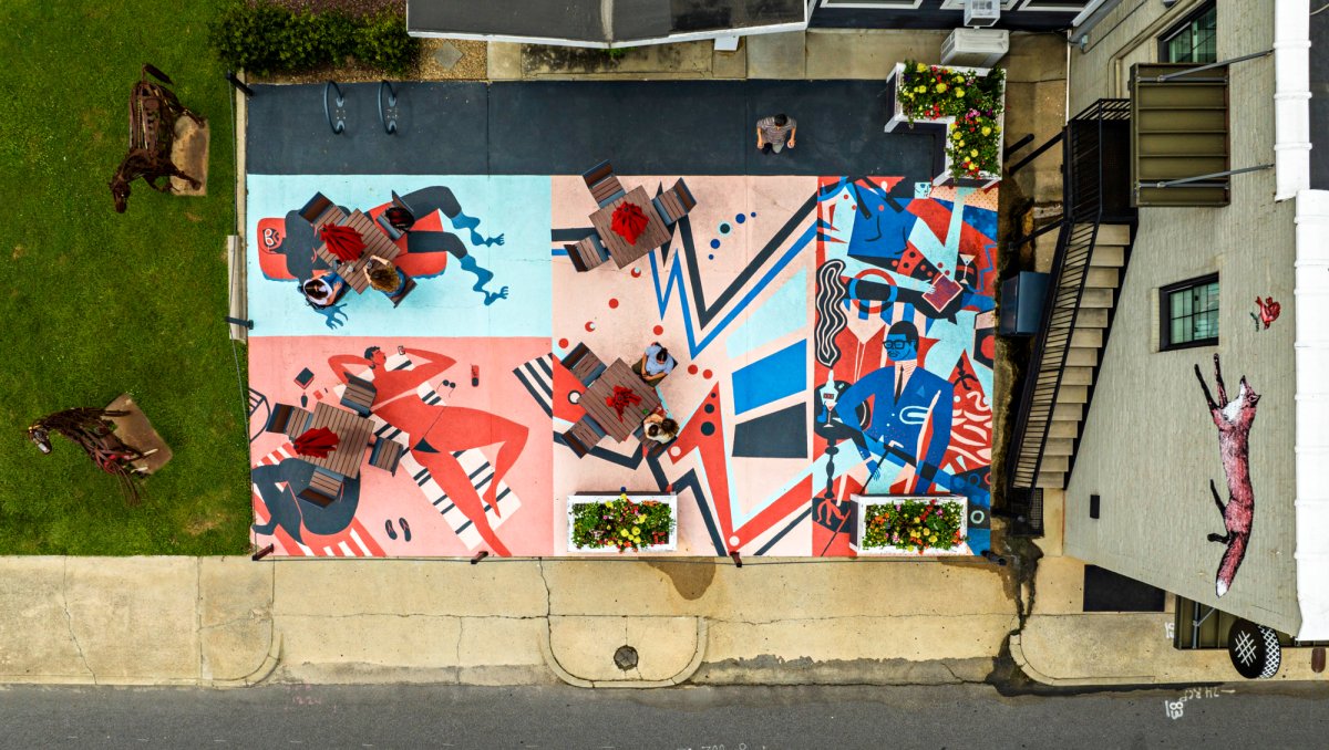 Aerial view of a vibrant outdoor patio space featuring colorful geometric and figurative murals on the ground. The murals depict abstract human figures and bold patterns in shades of blue, red, pink, and white. Wooden tables with red umbrellas are spaced across the artwork, with a few people seated. To the left, there's green grass with sculptures of horses, and on the right, a white brick building displays a painted fox on its exterior wall. A two-lane street borders the scene at the bottom of the image.