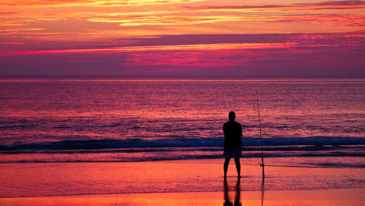 Silhouette of man surf-fishing on beach with bright pink and orange sunset