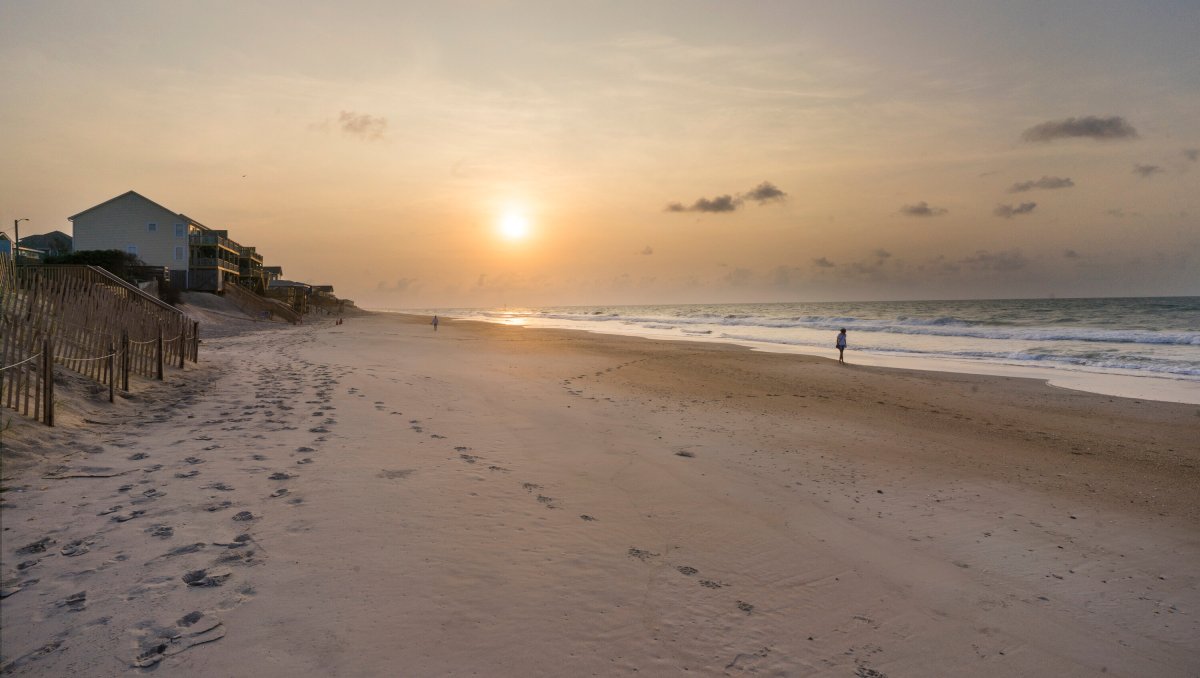 Sun rising in distance over wide-open beach with a few people walking on sand and rentals to left