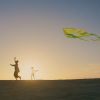 backlit woman and young girl with arms in air fly green triangle-shaped kite with tails on sand at sunset