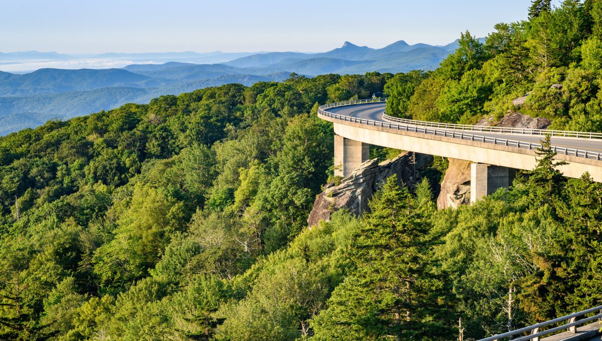 Linn Cove Viaduct to the right with green trees to the left and mountain ridges in background on clear day