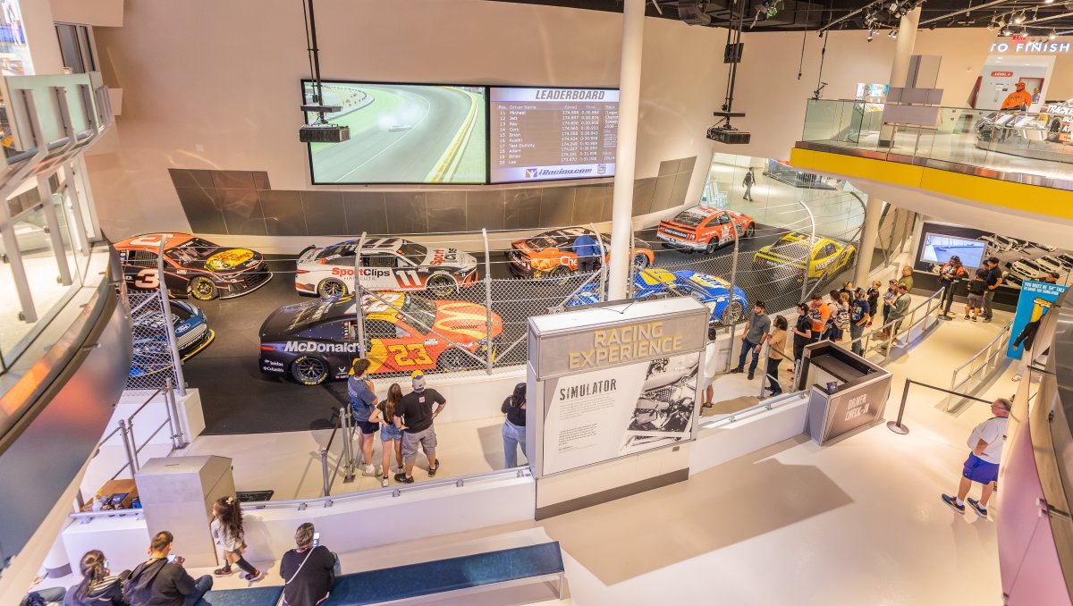 Elevated side view of the NASCAR Cup Series cars available for simulation racing with visitors lined up waiting their turn.