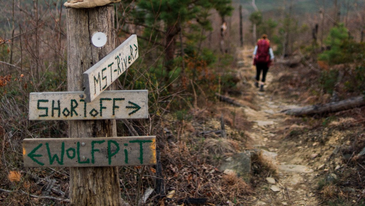 Person walking down trail with sign in foreground pointing to various mountains