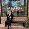 Woman sits on a bench outside Venture Chocolate & Wine Co., holding a drink, smiling and enjoying the cozy ambiance.