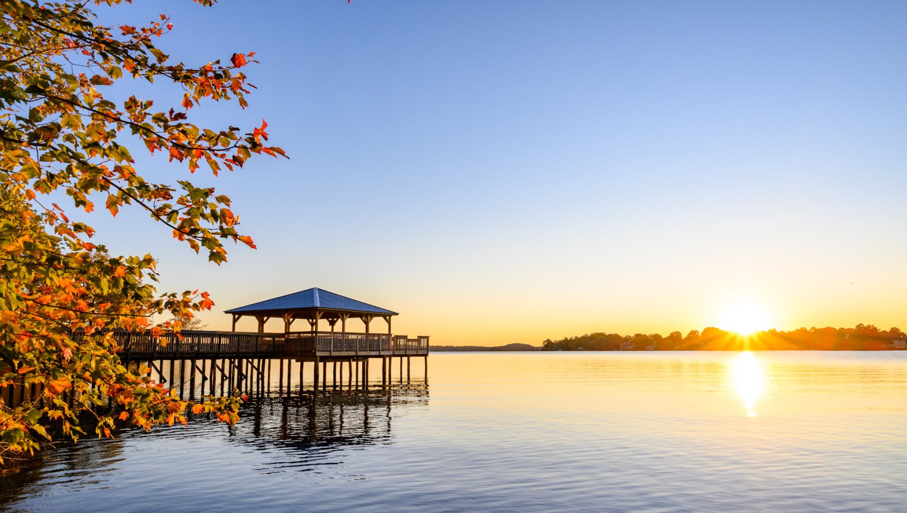 Pier on Lake Norman with sun setting behind trees on horizon in background with fall foliage in foreground