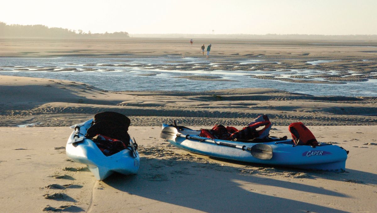 Two blue kayaks on sand in forefront with three people exploring sand and water on Bear Island during daytime