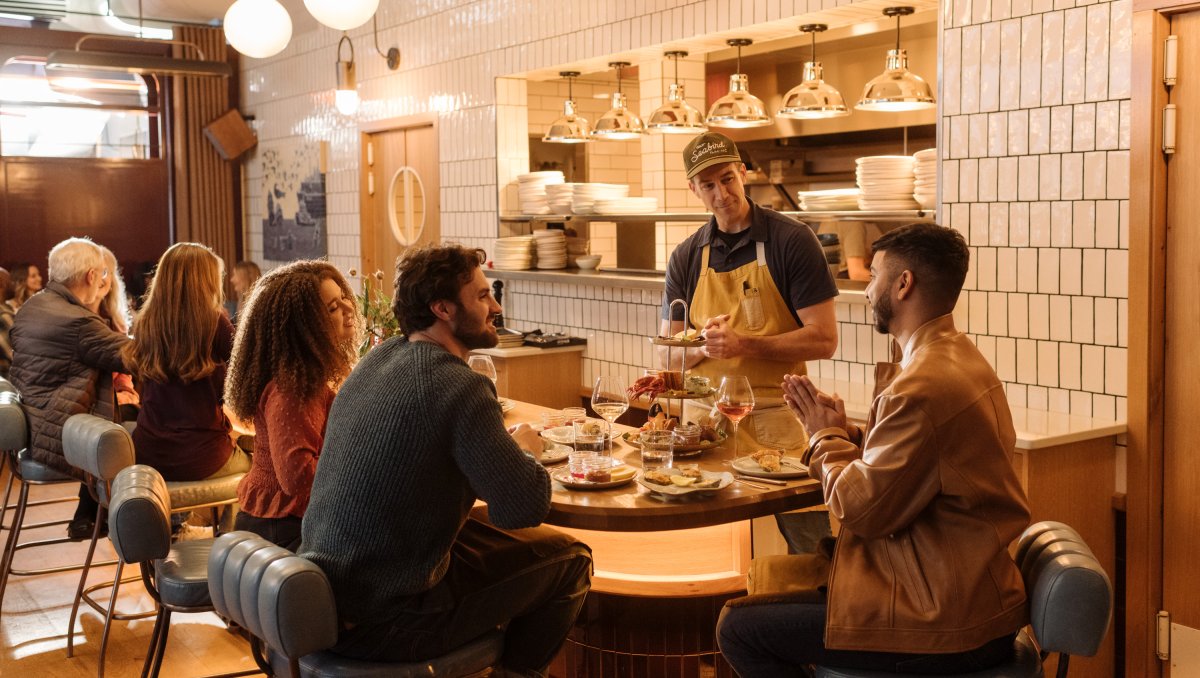 Chef chatting with diners at seafood restaurant with kitchen and other diners in background.