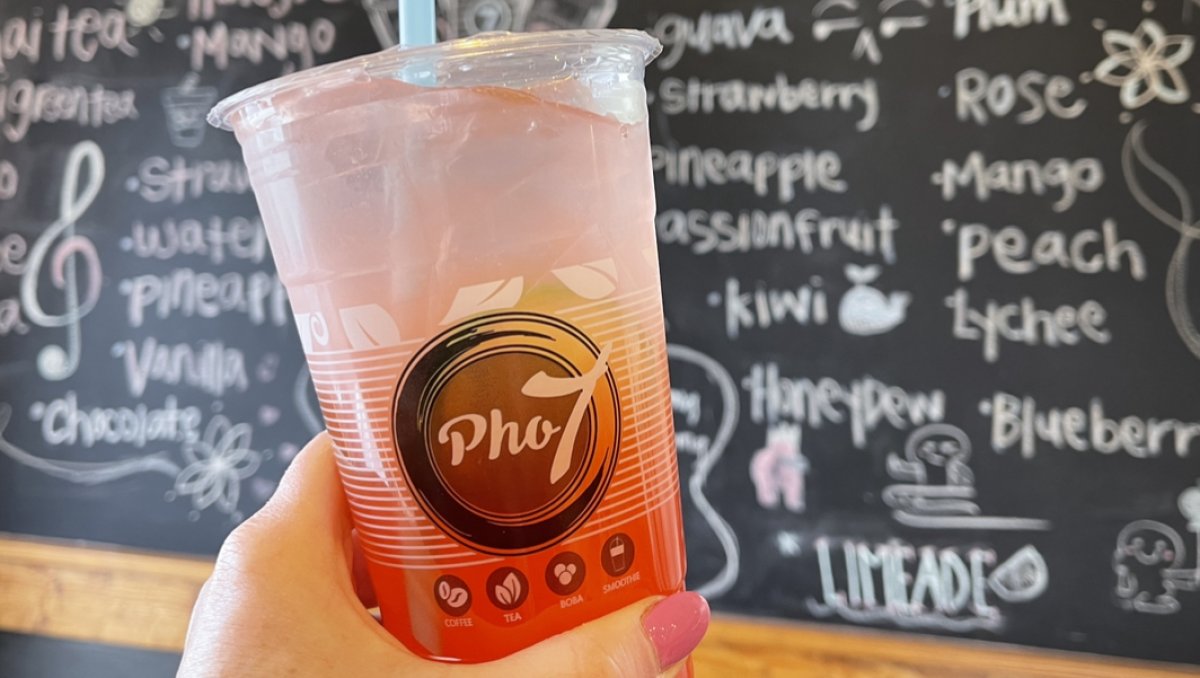 Person holding pink bubble tea in front of handwritten menu of drinks