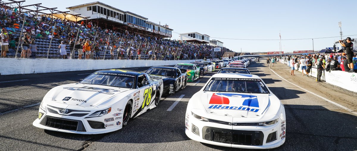 Two lines of race cars on a track with stands filled with people to the left