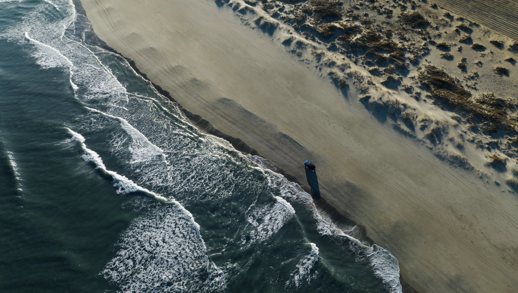 Aerial view of 4x4 vehicle driving on sand alongside ocean.