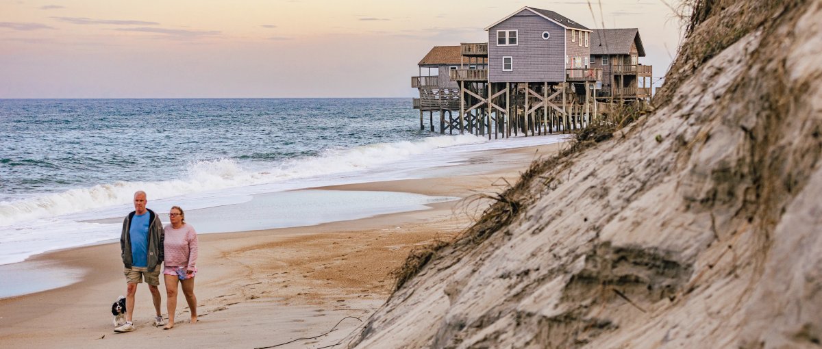 Couple walking dog along beach with ocean washing under house in background