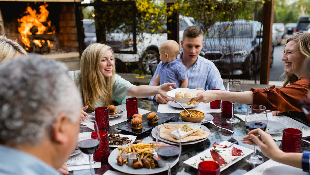 Family sitting around table sharing plates outside with fire roaring in fireplace in background