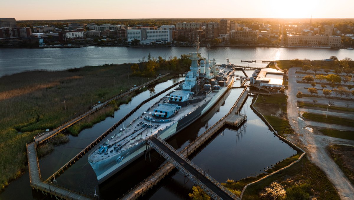 Aerial shot of Battleship North Carolina in river with city in background during daytime.