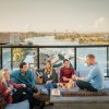 Friends sitting on bar rooftop couches enjoying drinks with Wilmington and river in background.