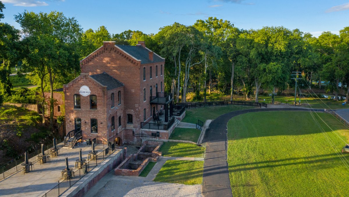 Aerial of brick distillery with green space in front surrounded by green trees on sunny day.